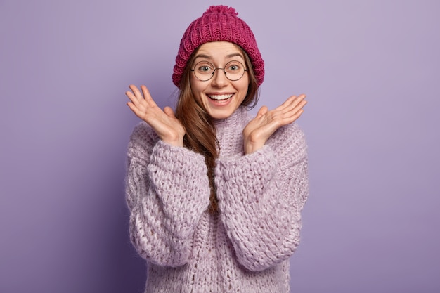 Foto de uma mulher europeia emotiva e feliz gesticula com as duas mãos, aperta as palmas das mãos, usa óculos redondos, vestida com um chapéu de inverno, jumper de malha, isolado sobre a parede roxa Conceito de boas emoções