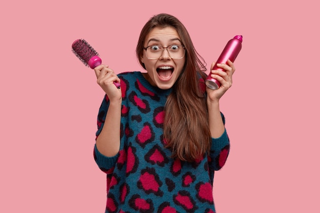 Foto de uma mulher europeia surpresa com a boca bem aberta em uma expressão positiva, segurando o spray e a escova de cabelo e olhando para o espelho radiante