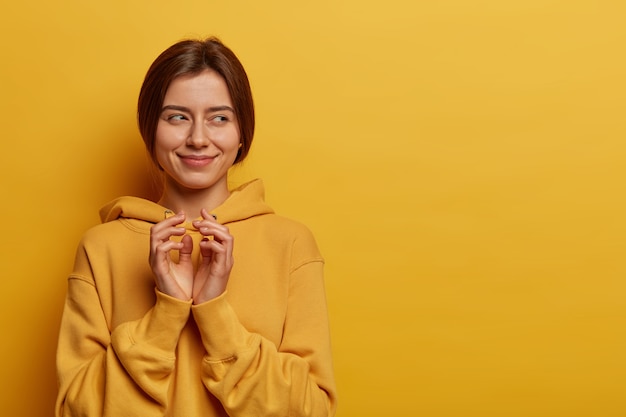 Foto de uma mulher feliz e misteriosa inclina os dedos e tem uma intenção misteriosa, trama alguma coisa, tem um olhar cunny de lado, usa um moletom, posa sobre a parede amarela, copie a área de espaço para seu texto.