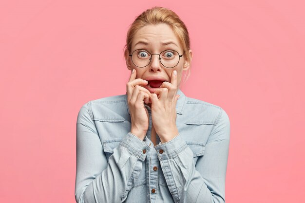 Foto de uma mulher loira de olhos azuis com aparência nervosa, mantém os dedos na boca, olha para a câmera, fica preocupada com medo de ir ao dentista, usa uma jaqueta jeans da moda,