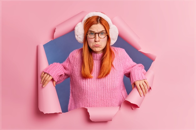 Foto de uma mulher ruiva decepcionada e frustrada franze os lábios e parece tristemente insatisfeita ao ouvir palavras ofensivas e usa um suéter de malha com protetores de ouvido quente.