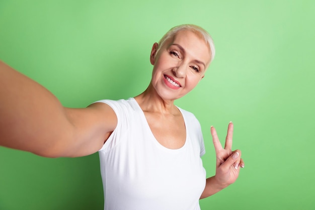 Foto de uma mulher sênior atraente atirar selfie mostra os dedos paz legal v-símbolo isolado sobre o fundo de cor verde.