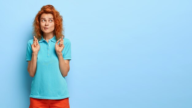 Foto de uma mulher séria de cabelos ruivos franze os lábios, mantém os dedos cruzados, ora pelo sucesso, usa camiseta azul, faz sinal de oração