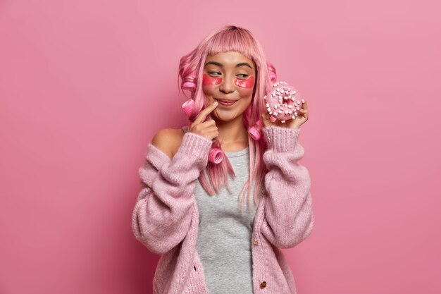 Foto de uma mulher sorridente, parecendo um goog, tem um dente doce e parece com apetite em um delicioso donut, usa rolos de cabelo, tem um penteado rosa