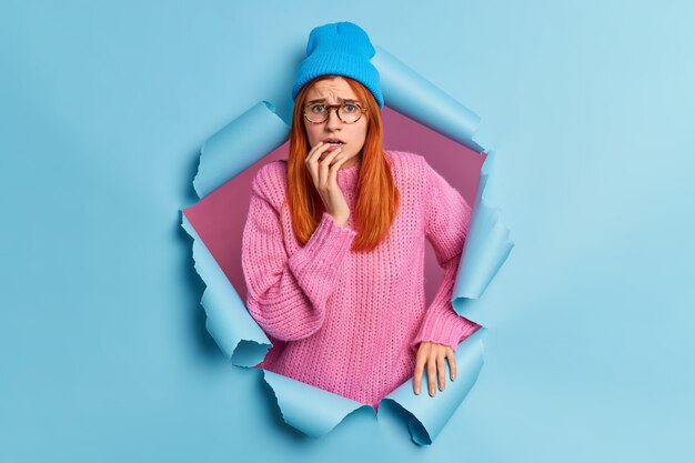 Foto de uma ruiva preocupada, mulher milenar, parece assustada, fica ansiosa usa um suéter de malha azul com um chapéu que rompe o buraco do papel