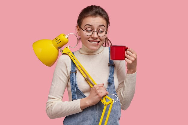 Foto de uma senhora de aparência agradável olhando com alegria para uma xícara de café, usando óculos redondos e carregando uma luminária de mesa amarela