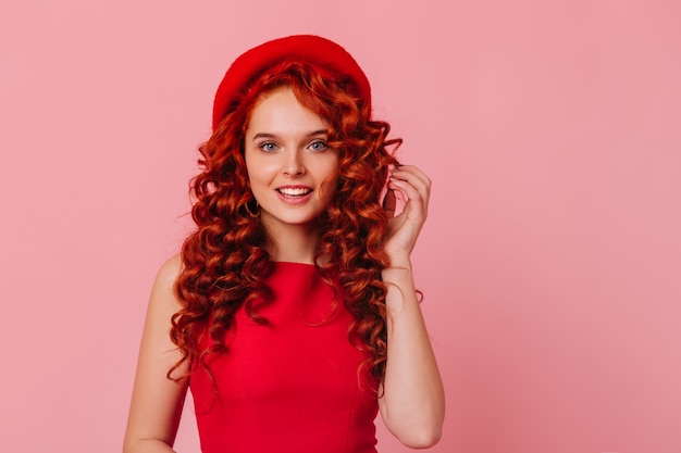 Foto de uma senhora positiva com olhos azuis e cachos vermelhos no espaço rosa. garota com cocar brilhante e top vermelho, olhando para a câmera.