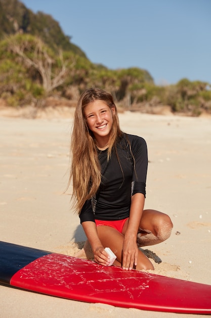 Foto de uma surfista ativa vestida de maiô, tem cabelos longos, sorriso agradável no rosto, prepara sua prancha de surf encerando a superfície antes da sessão