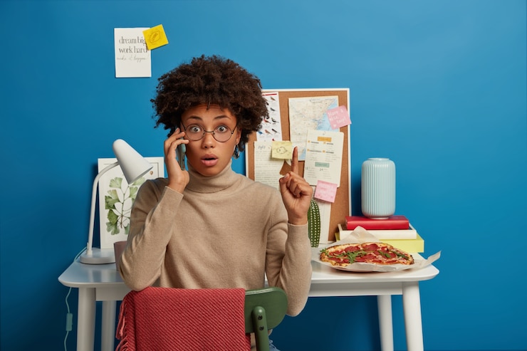 Foto grátis foto de uma trabalhadora chocada e surpresa de óculos aponta para cima durante uma conversa telefônica, liga para alguém via smartphone, senta-se na cadeira contra a mesa com pizza, quadro com notas, abajur, parede azul