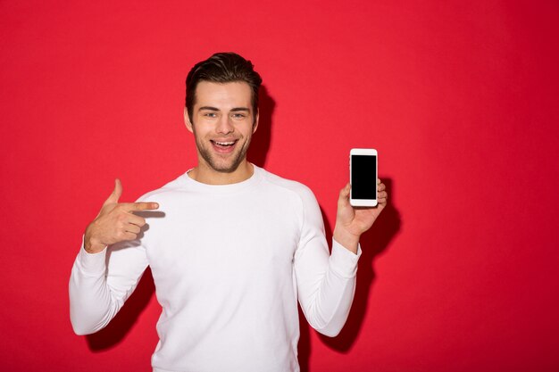 Foto do homem feliz na camisola olhando enquanto segura o smartphone e apontando para ele sobre parede vermelha