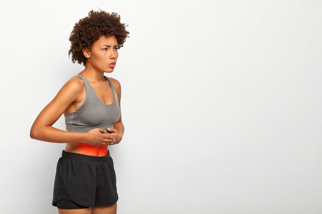 Foto do perfil de uma modelo afro-americana que sofre de dor de estômago, tem dor abdominal, toca a barriga, usa top e shorts, franze a testa por causa de sensações desagradáveis, poses contra um fundo branco