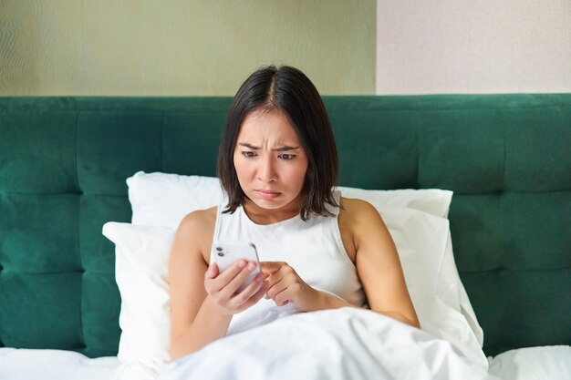 Foto do quarto de uma mulher asiática deitada na cama parecendo assustada e chocada com a tela do smartphone lendo a mensagem com o rosto preocupado