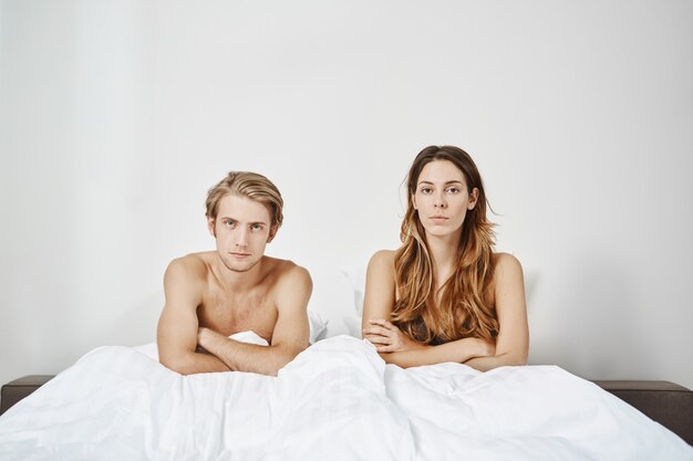 Foto do quarto do casal sentado na cama nua sob o cobertor com as mãos cruzadas e expressão insatisfeita. Jovens adultos em relacionamento têm problemas na vida sexual que não desejam discutir