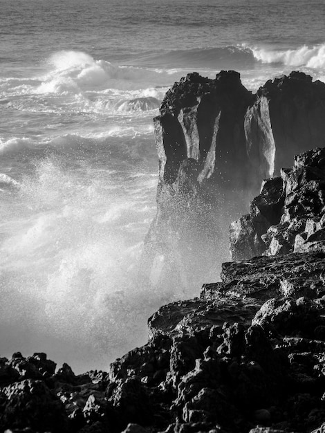 Foto em escala de cinza de ondas fortes atingindo grandes rochas em uma costa com respingos de água