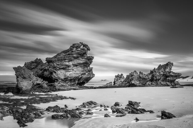 Foto em escala de cinza de rochas na praia com céu nublado