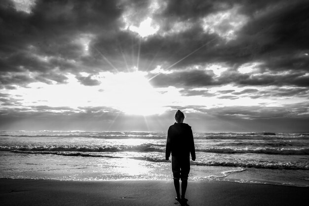 Foto em escala de cinza de uma mulher em pé na praia com a luz do sol no céu nublado