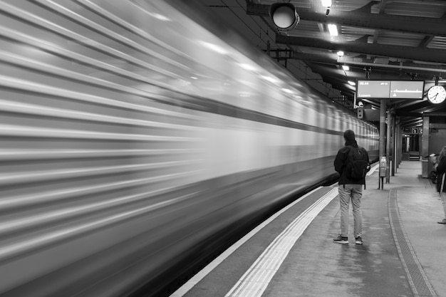 Foto em tons de cinza de um homem esperando um trem na estação e um trem borrado em movimento