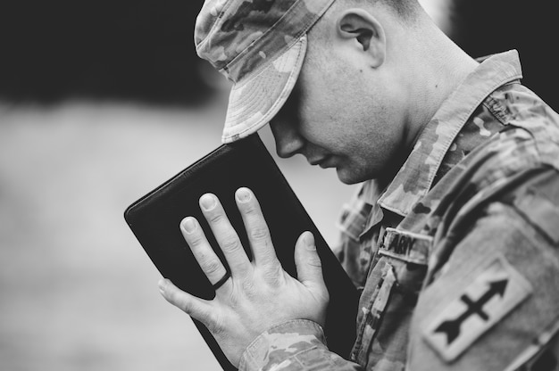 Foto em tons de cinza de um jovem soldado orando enquanto segura a bíblia