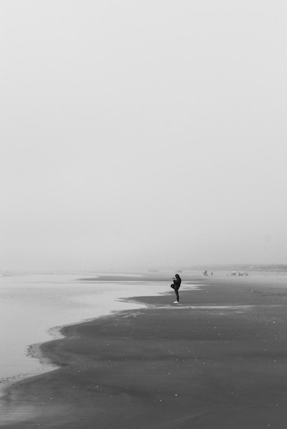 Foto em tons de cinza de uma pessoa solitária caminhando na praia sob nuvens escuras