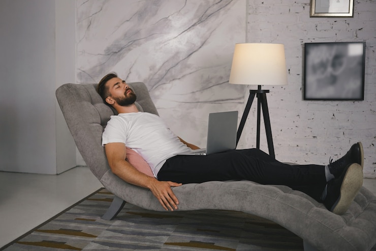 Foto grátis foto espontânea de um jovem com a barba por fazer em sapatos, deitado confortavelmente em um sofá cinza com um computador portátil no colo, tirando uma soneca ou meditando, mantendo os olhos fechados, ouvindo música relaxante