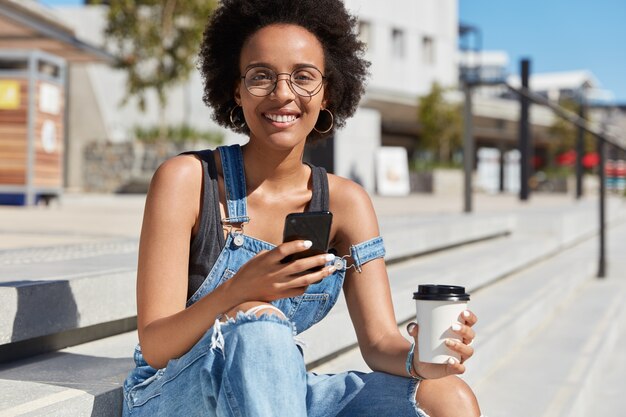 Foto externa de blogueiro relaxado e positivo com pele escura, bate papo online, usa a conexão gratuita à Internet na cidade, gosta de café aromático para viagem, usa óculos e macacão, posa em escadas com bebidas