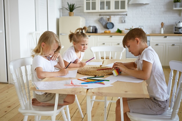 Foto franca de três adoráveis crianças, irmãos de aparência europeia, sentados à mesa da cozinha e fazendo desenhos de família juntos, usando lápis coloridos, concentrando-se em expressões sérias