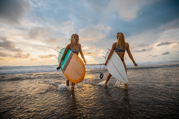 Foto grande angular de duas mulheres caminhando na praia com pranchas de surfe