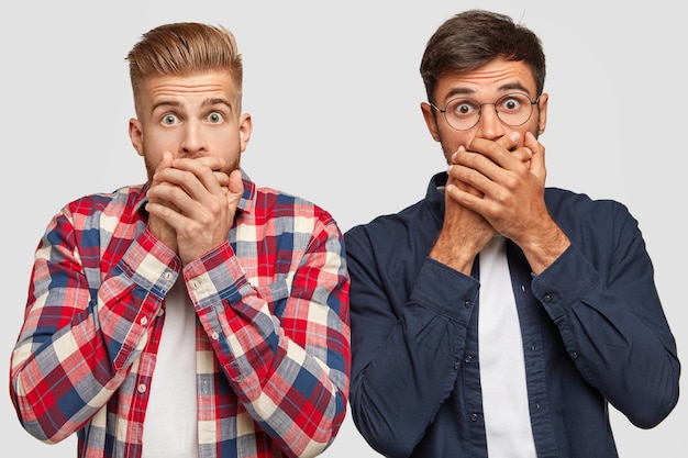 Foto horizontal de caras elegantes ou colegas de trabalho surpresos mantendo as mãos na boca, olhando para a câmera, sentindo espanto, ficando próximos um do outro