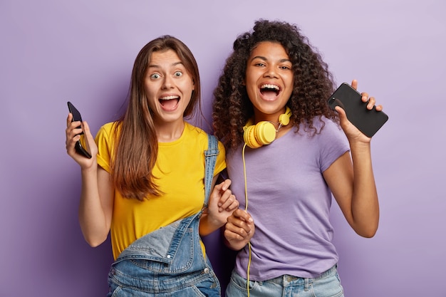 Foto horizontal de melhores amigos felizes se encontrando durante o fim de semana, divirta-se com tecnologias modernas, dance ao som da música, olhe com alegria para a câmera