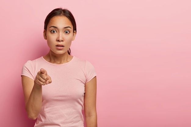 Foto horizontal de mulher asiática assustada aponta o dedo indicador, fica com vergonha de notar algo estranho, tem cabelo escuro penteado em rabo de cavalo, usa uma camiseta rosada, isolado na parede rosa