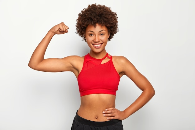 Foto horizontal de mulher de pele escura positiva mostra o bíceps, demonstra mão forte, tem corpo esguio, usa sutiã esportivo, sorri agradavelmente, isolado sobre fundo branco.