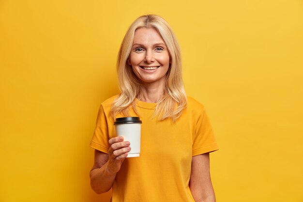 Foto horizontal de mulher europeia loira com sorriso agradável, maquiagem mínima segura xícara descartável de café vestida com camiseta casual