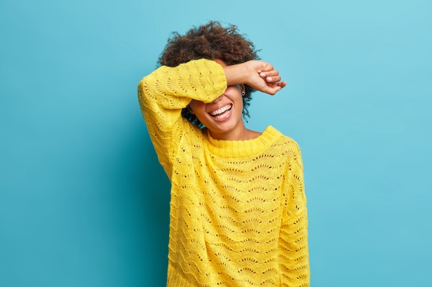 Foto horizontal de mulher positiva de cabelo encaracolado ri sinceramente e cobre os olhos com o braço rindo de uma piada engraçada vestida com um macacão de malha amarelo isolado na parede azul