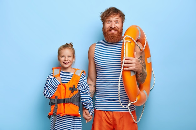 Foto horizontal de pai e filho felizes em um colete salva-vidas protetor, carregue salva-vidas, passe as férias de verão juntos, aprenda a nadar, expresse boas emoções