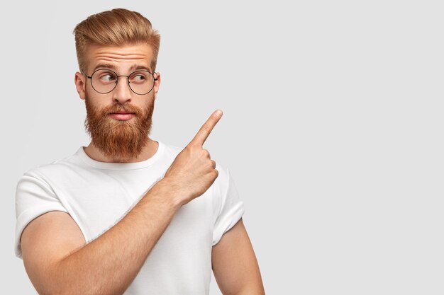 Foto horizontal de um atraente homem branco com barba e cabelo ruivos, apontando com o dedo da frente de lado, vestindo uma camiseta em uma cor com a parede