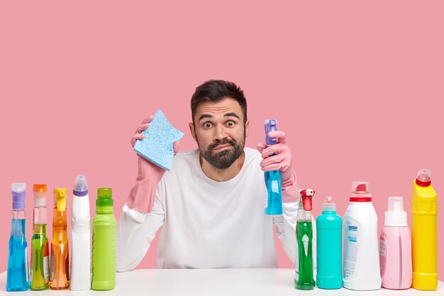 Foto horizontal de um cara barbudo do serviço de limpeza segura a esponja e o spray de limpeza, limpa os móveis da cozinha com um pano, usa diferentes solventes