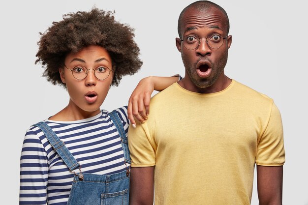 Foto horizontal de um casal de família de pele escura e emotivo que não espera receber contas, fica de queixo caído, não vê como sair, não tem dinheiro para pagar, isolado sobre uma parede branca. Conceito Omg