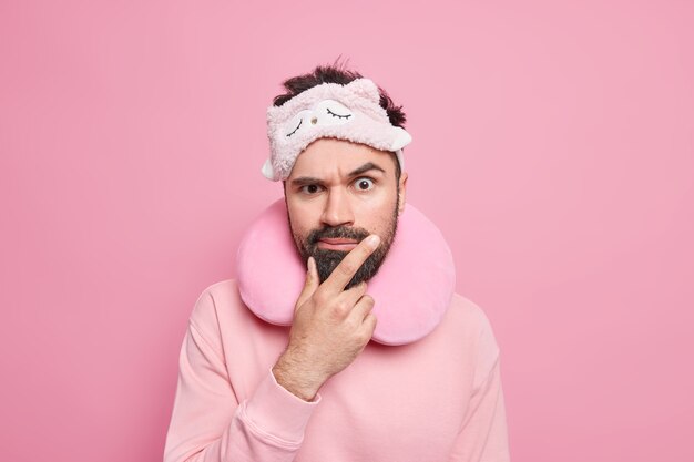 Foto horizontal de um homem adulto barbudo sério com expressão rígida, usando um travesseiro de viagem confortável e uma máscara de dormir vestida casualmente