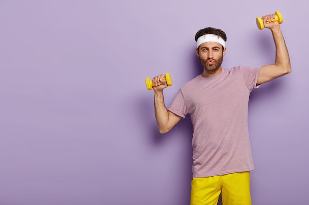 Foto horizontal de um homem bonito com a barba por fazer malhando na academia, treina bíceps com o instrutor esportivo, usa roupa ativa, bandana branca