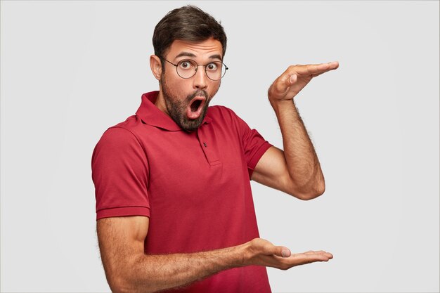 Foto horizontal de um homem branco surpreso e bonito em uma camiseta vermelha, mostra a altura de algo, abre a boca com admiração