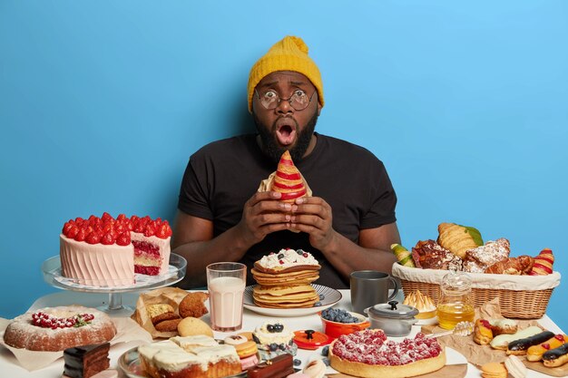 Foto horizontal de um homem com a barba por fazer segura um croissant, tem uma expressão facial estupefata, choque por ter vício em açúcar, usa chapéu amarelo, camiseta e óculos, olha em estupor.