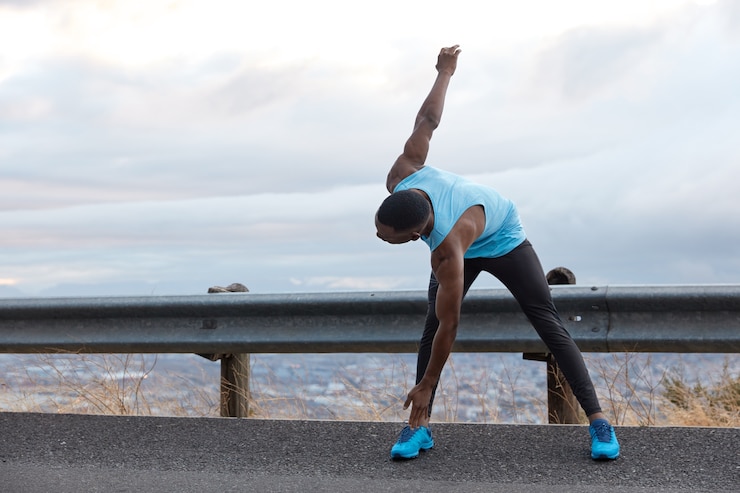 Foto grátis foto horizontal de um homem de pele escura se inclina para os pés, faz exercícios de alongamento, tem formato de corpo musculoso, fica no asfalto sobre um céu azul claro com espaço de cópia para seu conteúdo de publicidade, texto