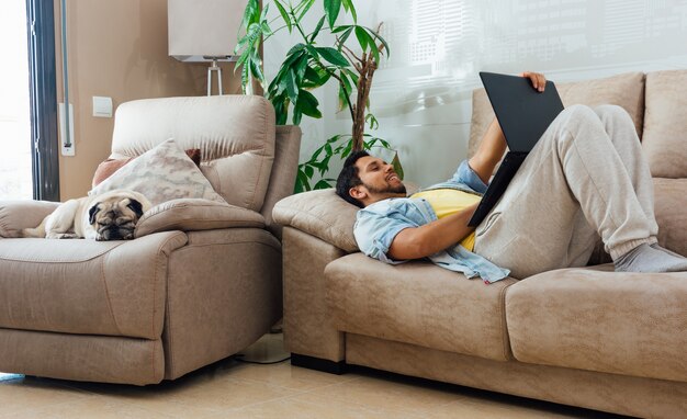 Foto horizontal de um homem deitado no sofá em casa e trabalhando com um laptop preto