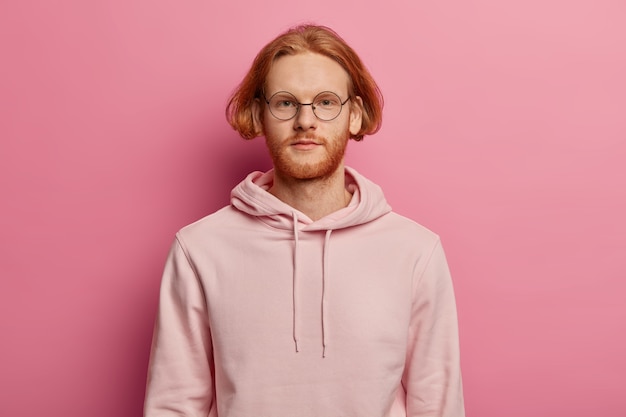 Foto horizontal de um homem europeu barbudo de aparência séria olha diretamente, tem uma expressão calma, usa um moletom casual, posa contra uma parede rosada, fala com alguém, tem um penteado bob