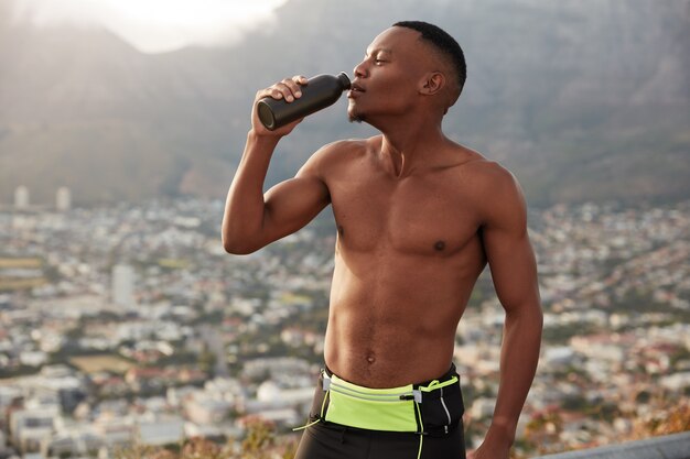 Foto horizontal de um homem negro com corpo esportivo, hidrata-se com água, segura a garrafa, sente sede após o treino cardiovascular, respira por causa de um ataque cardíaco, sente-se desidratado, se opõe à paisagem de montanha