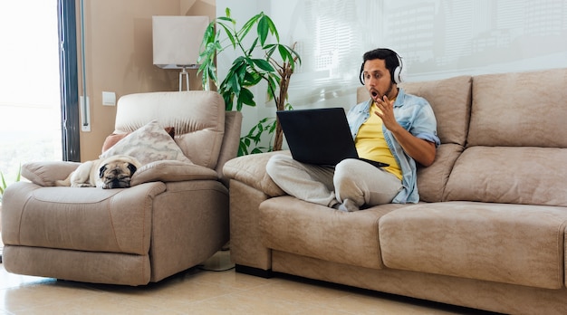 Foto horizontal de um homem sentado no sofá, trabalhando com um laptop e se sentindo chocado