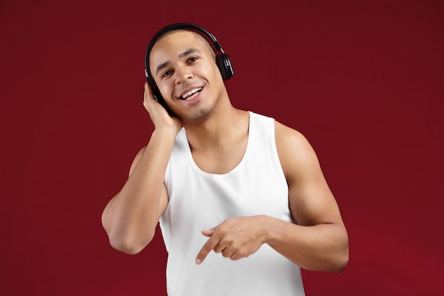 Foto horizontal de um jovem afro-americano simpático e bonito em uma elegante camisa sem mangas, sorrindo alegremente e apontando o dedo enquanto ouve música em fones de ouvido, cantando junto com uma melodia