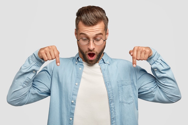 Foto horizontal de um jovem com barba por fazer surpreso aponta para baixo, abre a boca amplamente, vê algo impressionante no chão, usa uma camisa elegante, isolada sobre uma parede branca. conceito de pessoas e emoções