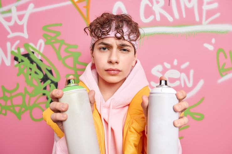 Foto grátis foto horizontal de um rapaz infeliz vestido com um moletom segurando duas garrafas de aerossol fazendo grafite criativo na parede da rua com expressão insatisfeita