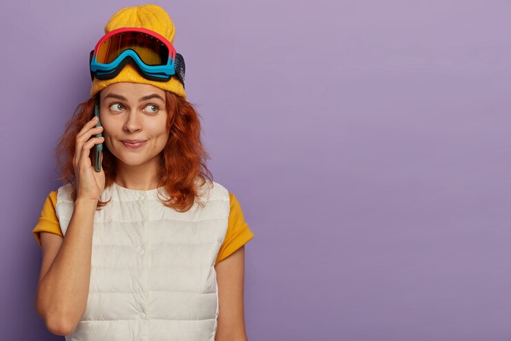 Foto grátis foto horizontal de uma adorável jovem ruiva em uma estação de esqui, faz um telefonema, usa um chapéu amarelo e colete branco, fica em uma parede roxa